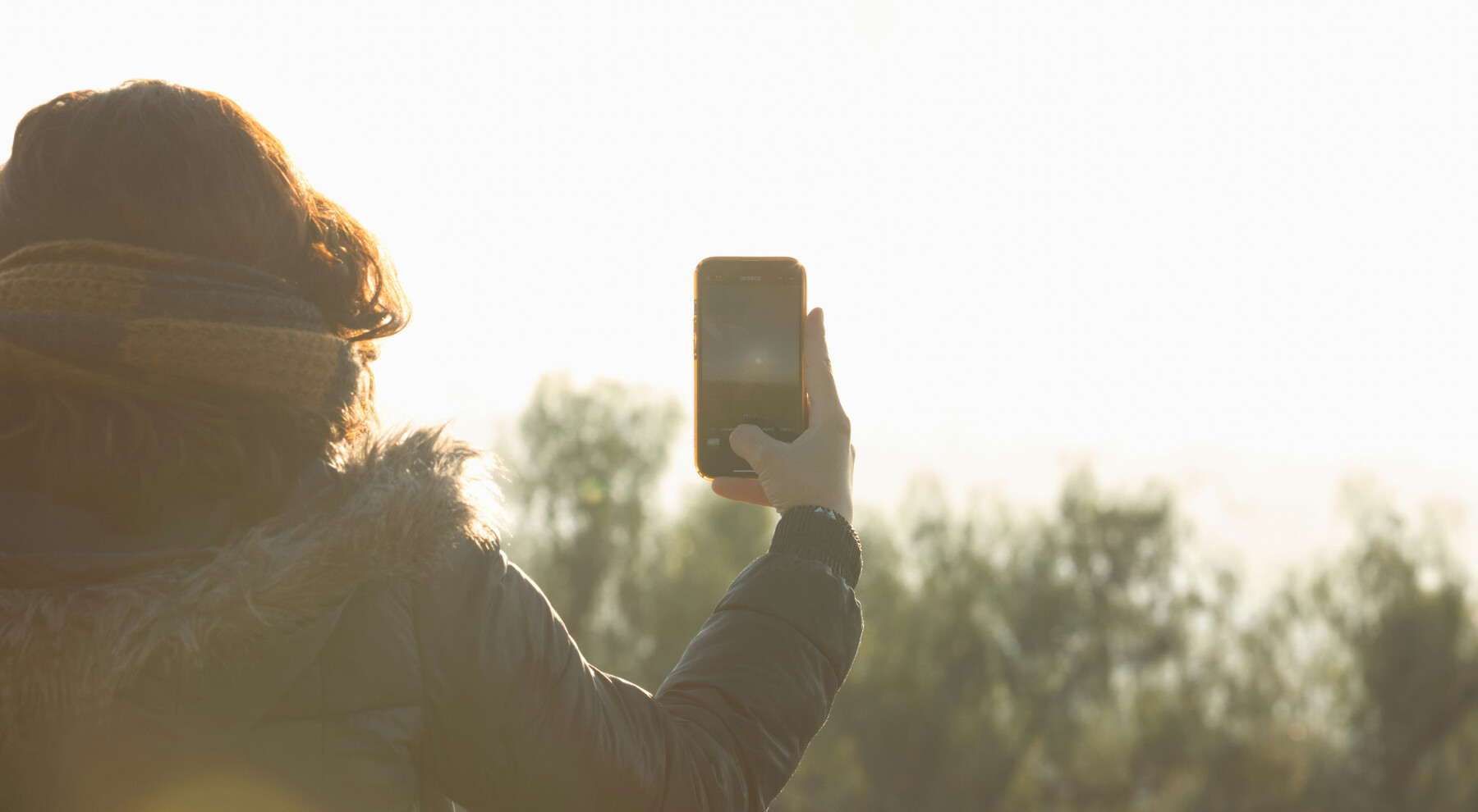 Egy hölgy a tájat fotózza a mobiltelefonjával, miközben gyönyörűen süt a nap.